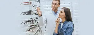 A man and a woman are standing in front of a glass shelf with eyeglasses, the man is pointing at something and the woman is looking at it with her hand on her chin.