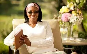 A woman wearing a white shirt and sunglasses sits on a chair in an outdoor garden, holding a book and smiling for a photo.
