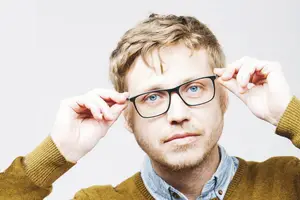 A man with short brown hair and blue eyes wearing glasses and a light brown sweater.