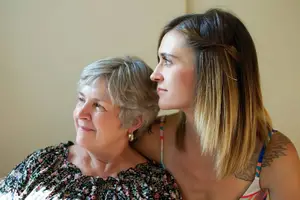 An elderly woman with gray hair is sitting on a couch, and a young woman with blonde hair is looking at her, and the elderly woman is wearing a floral dress, and the young woman is wearing a sleeveless dress with a tattoo on her shoulder, and they are both looking at each other.