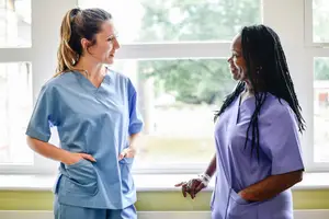 Two nurses in blue scrubs are standing in front of a window, smiling and looking at each other.