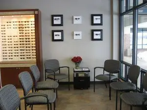 An interior of a room that has a display case with glasses and a table with a flower vase on top, a cabinet, and chairs. It also has picture frames and glass windows. The room is probably a clinic.