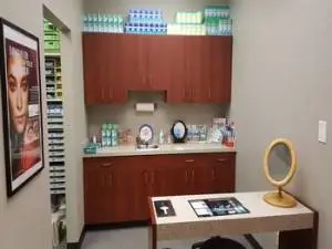 A well-organized and clean bathroom with wooden cabinets, a countertop, a sink, and a mirror. A poster is mounted on the wall, and there is a roll of paper towels on the countertop. Various items are on the countertop, including a bottle, a bowl, and a plate. A shelf is on the left side, and there are bottles and other items on it.