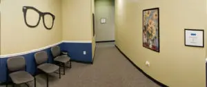 A view of a hallway with yellow walls, a blue border, and a few chairs on the left. A pair of glasses is mounted on the yellow wall. A picture frame is mounted on the wall, and a power outlet is on the wall to the left. A small window is on the wall in the hallway. The hallway has a carpeted floor.