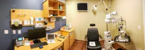 A medical office features a desk with a computer, a telephone, and various equipment, including an eye exam machine and a monitor mounted on the wall.