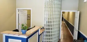 A marble counter with a blue flower vase stands in front of a glass block column. Behind it, a hallway leads to a room with a picture frame and an open door. The walls are painted in beige and blue, and there is a white ceiling light.