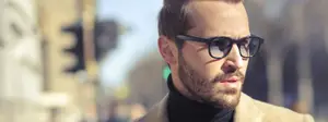 Man with beard wearing black glasses and brown coat standing on a city street.