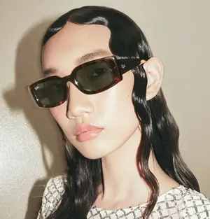 A woman with long black hair wearing brown-tinted sunglasses and a white patterned dress poses indoors.