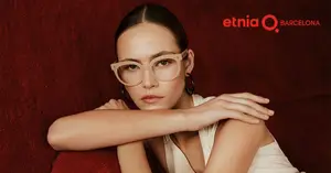 A woman wearing glasses and a white dress leaning against a red wall with the Etnia Barcelona logo
