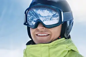 A smiling person wearing a helmet and goggles is standing in a snowy area, possibly posing for a photo.