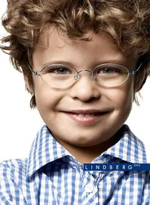 A young boy with curly hair wearing a blue and white checkered shirt and clear glasses is smiling for the camera.