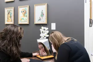 Two women are looking at a mirror on a desk, one of them is holding her cheeks, and there are several paintings on the wall behind them.