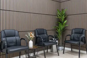 Three black leather chairs are arranged around a small table with a vase of flowers. The chairs are positioned against a wall with vertical stripes. The floor is wooden and the room is well-lit.