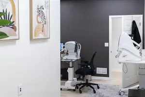 An optometrist's office with a desk, chairs, a machine, and a door leading to another room with a coat hanging on the door