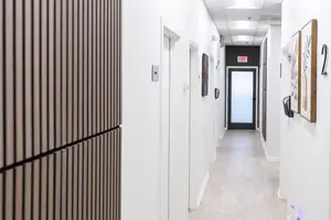 A corridor of an empty room with white walls and wooden floors, featuring a glass door and decorative framed pictures on the wall, with the number 2 visible above the door.