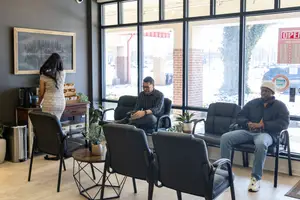 Three people are in a waiting room with glass walls, potted plants, and a desk with a coffee machine.