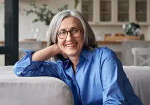 A smiling woman with gray hair wearing glasses and a blue shirt is sitting on a couch in a room with a kitchen in the background.