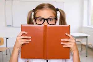 Girl with glasses hiding behind a book in a classroom