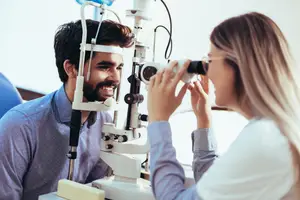 A man is looking at a machine while a woman is holding a device and looking at him in an eye clinic