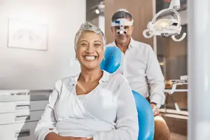 A woman with gray hair smiles at the camera in a dental clinic while a man wearing goggles stands behind her.