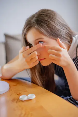 A young girl is looking at a white contact lens on a wooden table.
