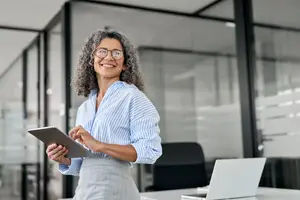 A smiling woman in glasses is standing in an office, holding a tablet, and standing near a desk with a laptop.