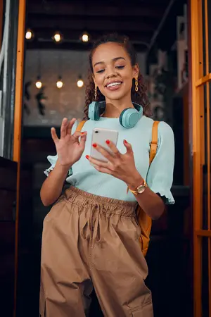 A woman with curly hair is standing in a dimly lit room, smiling, and looking at her phone with headphones on her neck.