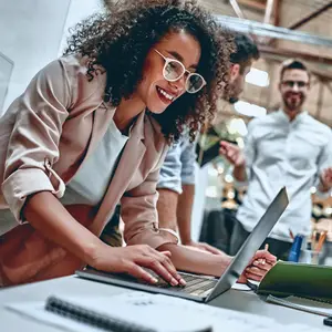 A woman wearing glasses is smiling while working on a laptop in an office with other people in the background.