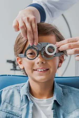 A young boy is sitting in a chair, wearing a pair of glasses, and seems to be getting an eye test by a doctor.