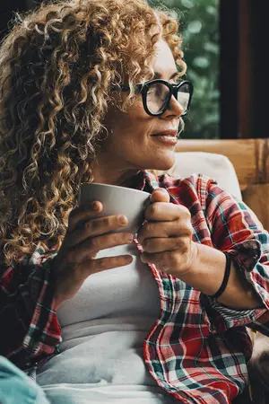 A woman wearing glasses, a plaid shirt, and a bracelet holds a white cup in her hands.