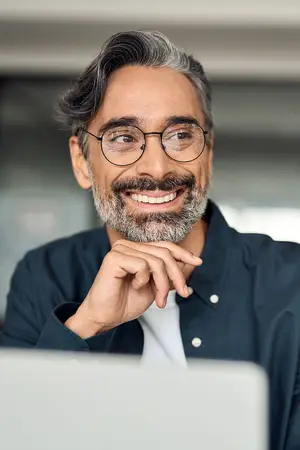 A smiling middle-aged man with gray hair and a beard wearing glasses and a navy blue shirt is looking at a laptop in an office setting.