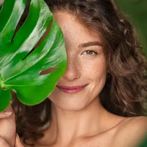 A smiling woman with curly hair and freckles is holding a large green leaf over her face in a green background.