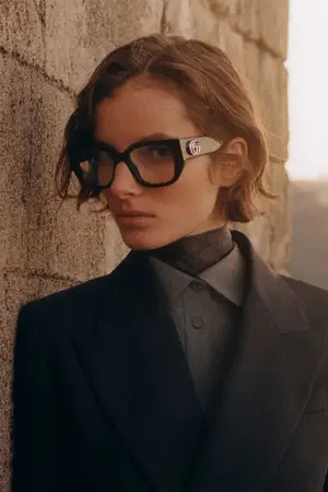 A woman wearing a black suit jacket, black turtleneck, and black glasses with silver accents stands next to a brick wall.