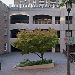 An image of a building with a parking area and a tree in front of it, along with a sign indicating no parking hours, a lamp post, and a staircase on the right side