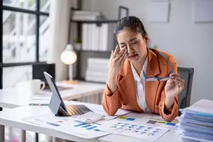 A woman is sitting at a desk in an office, rubbing her nose and looking at the computer screen. She is wearing glasses and an orange blazer. There is a calculator, papers, a cup, and a lamp on the desk. Behind her is a glass window, a shelf with books, and a whiteboard. There is a radiator on the floor next to the desk.