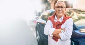An older man with glasses and a red scarf stands confidently next to a blue car on a sunny street.