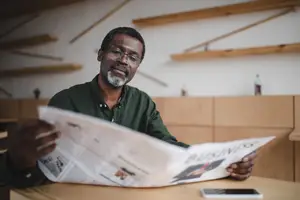 A man wearing glasses is sitting at a table and reading a newspaper