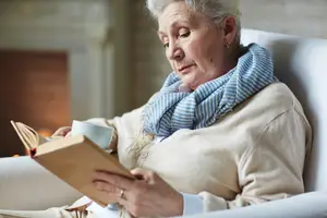 A senior woman is sitting on a white chair in a room and reading a book while holding a cup of coffee.