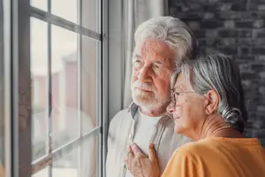 An elderly couple standing close to each other and looking out of a window.