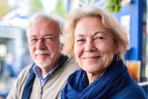 An elderly couple is smiling and looking at the camera while sitting in a restaurant. The woman is wearing a blue scarf and earrings. The man is wearing glasses. Behind them is a blurry view of a blue wall with a glass door and a potted plant.