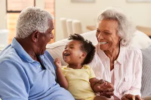 An elderly man and woman are sitting on a couch with a baby laughing in front of them.