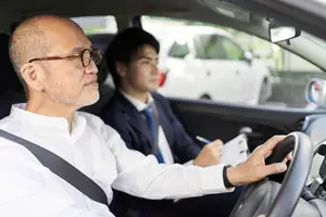 An older man wearing glasses drives a car with a younger man behind him holding a clipboard and writing