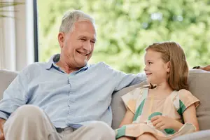 An elderly man and a young girl are sitting on a couch, smiling, with the man's arm around the girl's shoulder, near a window with trees outside