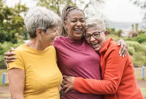 Three elderly women are laughing and hugging each other in a park.