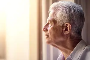 An elderly man with white hair is gazing outside a window with sunlight streaming in, wearing a white polo shirt.