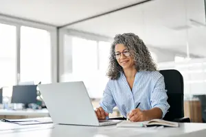 A woman sitting at a desk with a laptop and pen, smiling and holding a notebook and pen