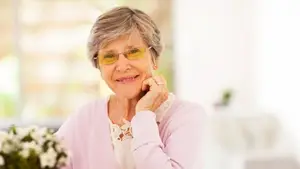 An elderly woman wearing glasses, smiling and touching her chin with her left hand, sitting in a room with a white wall and a flower in front of her.
