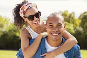 A man is giving a piggyback ride to a woman who is smiling and wearing sunglasses and a headband
