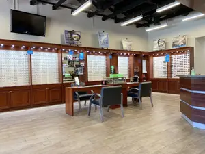 Interior of an optical shop featuring a wooden counter, a desk with chairs, a monitor, and various display cases with glasses, illuminated by ceiling lights and featuring posters on the wall.