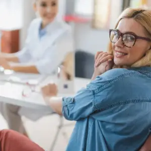 A woman wearing glasses and a blue shirt is sitting at a desk, smiling while looking at something. Another woman in a white lab coat is sitting in front of her, working on a computer.